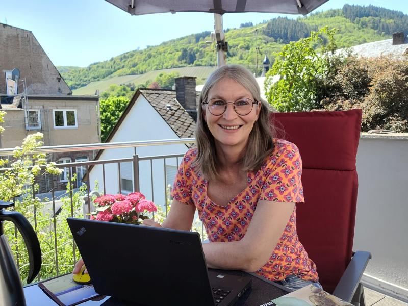 Frau auf Balkon unter Sonnenschirm lächelt in die Kamera, vor ihr ein Laptop, im Hintergrund Weinberge.