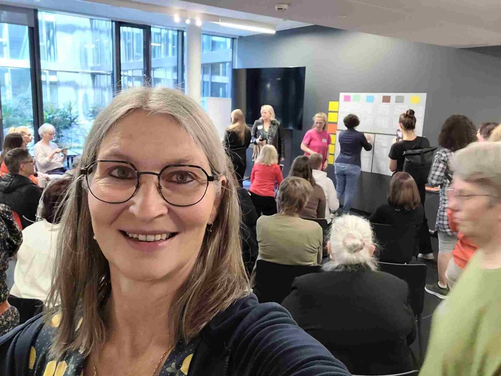 Selfie einer Frau in einem Raum mit vielen anderen, im Hintergrund eine Wand mit einem Whiteboard, an das Zettel geklebt werden.