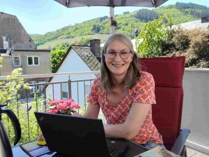 Frau lächelt in die Kamera, sitzt an Tisch auf Balkon vor Laptop, im Hintergrund Weinberge an der Mosel