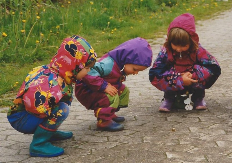 Drei kleine Kinder in Regenklamotten hocken auf einem Weg und beobachten eine Schnecke.