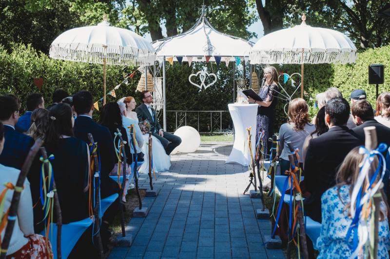 Freie Trauung in einem Garten mit Blick auf den Gang hin zum Brautpaar und der freien Rednerin unter Sonnenschirmen