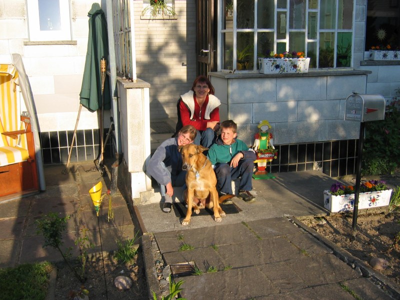 Frau mit zwei Kindern und Hund sitzt auf Treppe am Hauseingang in der Abendsonne