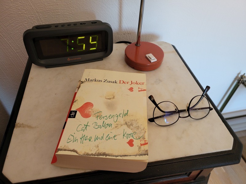 Buch "Der Joker" von Markus Zusak auf einem Nachttisch, daneben Brille und Wecker