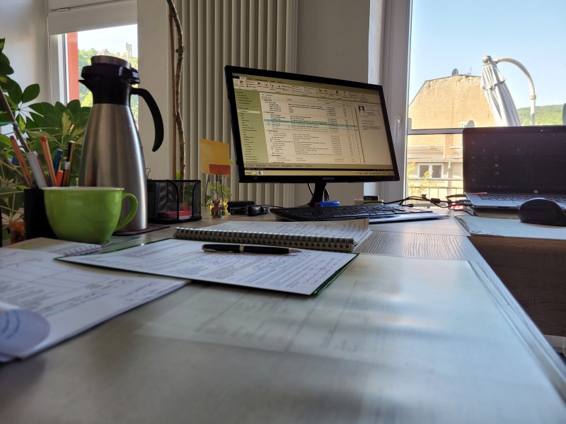 Ein Schreibtisch mit Bildschirm, Block, Thermoskanne und grüner Tasse und einem Blick aus einer Terrassentür