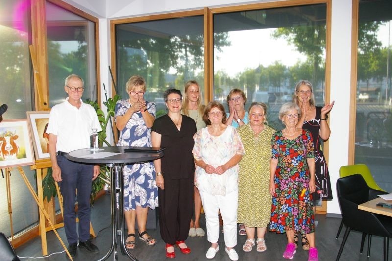 Neun Personen vor einer Fensterfront - Autor:innen der Lesung "Alle Neune - AusLeseZeit" am 4.7.25 in Wittlich
