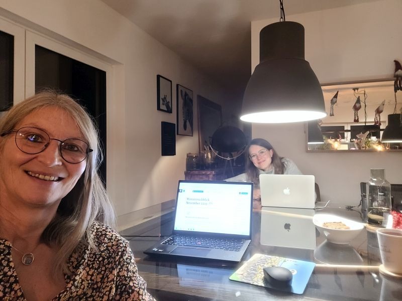 Selfie von zwei Frauen, die in an einem großen Tisch mit aufgeklappten Laptops sitzen.