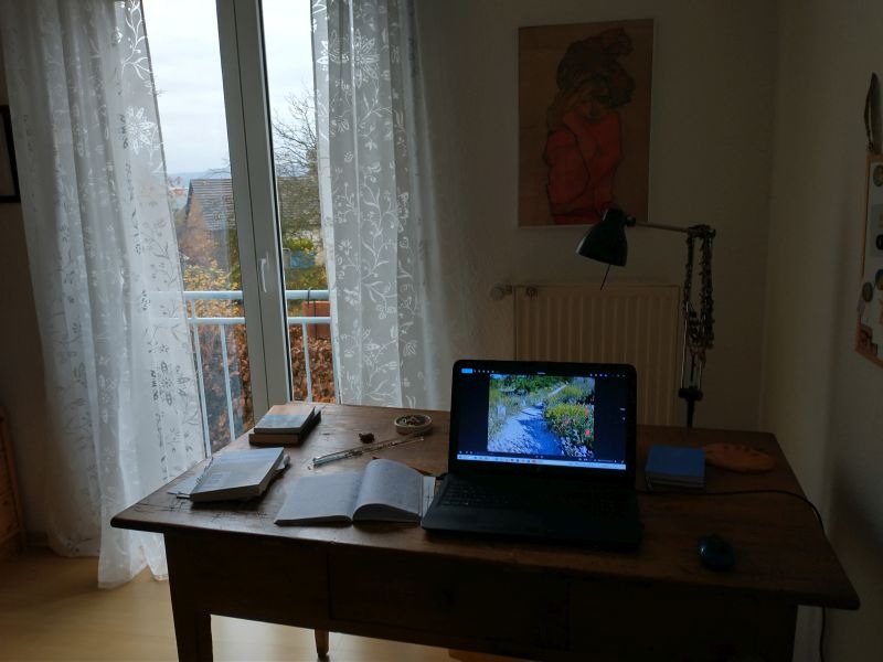 Schreibtisch mit Laptop und Notizbuch vor einem bodentiefen Balkonfenster