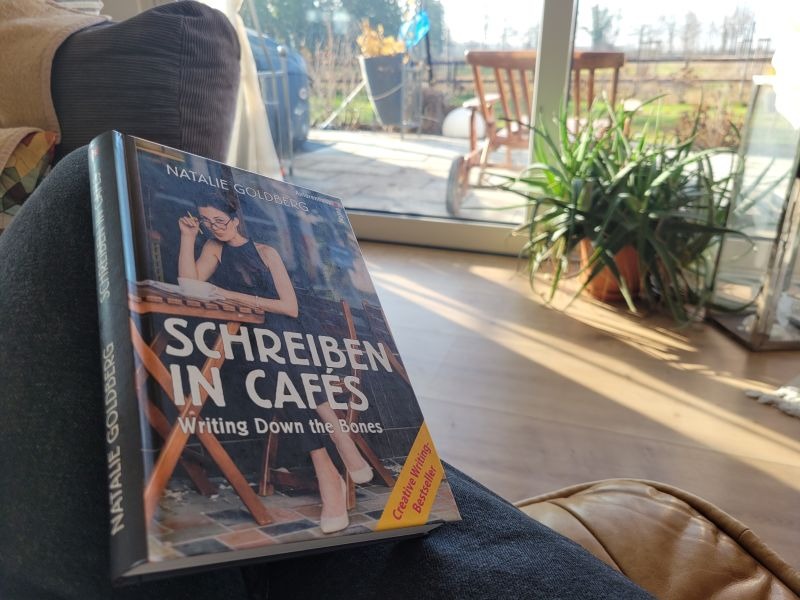 Das Buch "Schreiben in Cafés" von Natalie Goldberg liegt aufgeschlagen auf dem Knie der Leserin, Blick in einen Garten