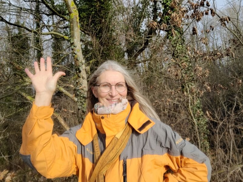 Selfie von Gabi Kremeskötter vor einem Wäldchen im Februar, sie hält ihre rechte Hand zum Gruße hoch, bekleidet ist sie mit einem orange-grauen Anorak.