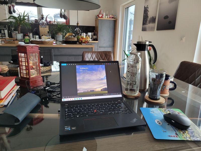 Laptop auf einem Tisch umgeben von Teekanne und Wasserflasche, im Hintergrund eine Küchenzeile.