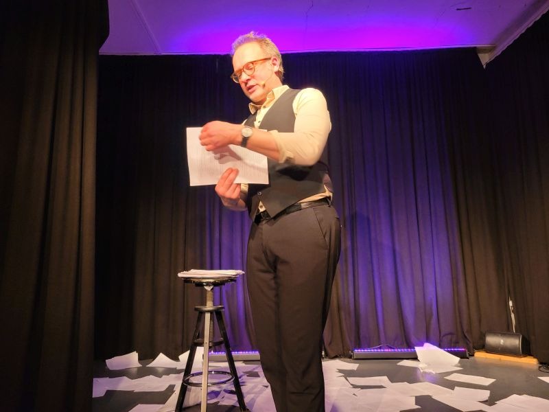 Kabarettit René Sydow auf der Bühne, hinter ihm zerstreute DIN-A-4 Blätter, neben ihm ein Barhocker