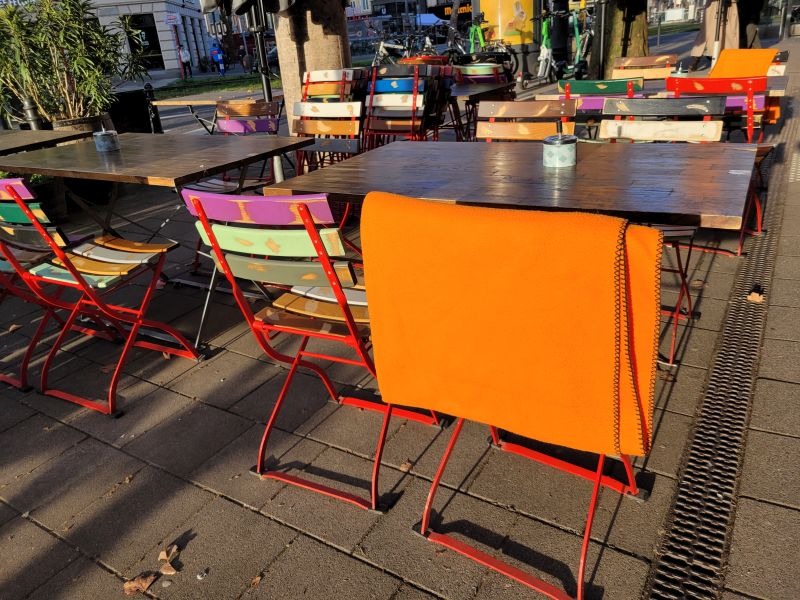 Biergartenstühle und Tische eines Straßencafés, über einer Lehne einee orangefarbene Fleecedecke
