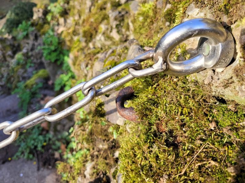 Eine Absperrkette, verankert in einer Natursteinmauer, glänzt in der Sonne.