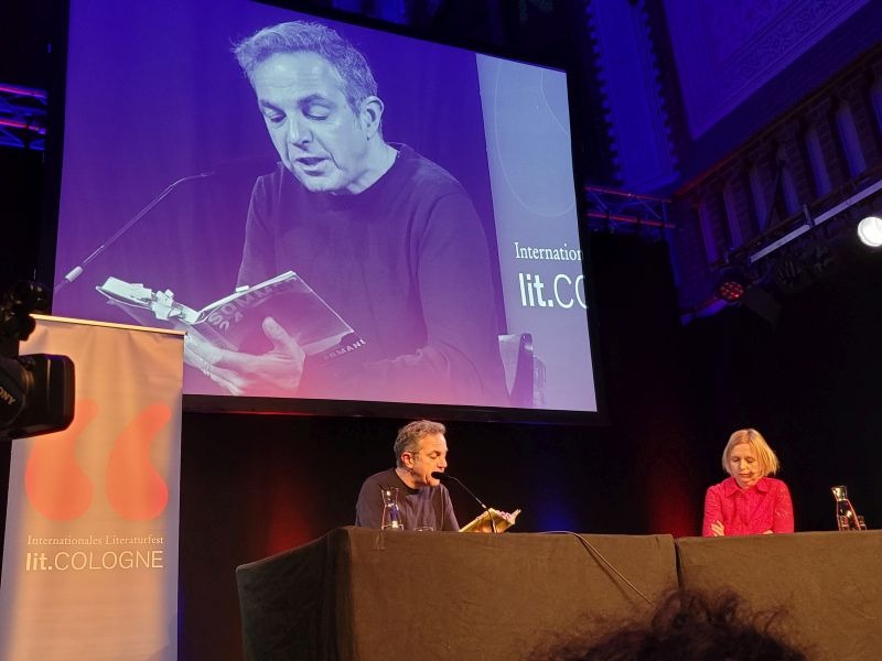 Navid Kermani auf seiner Lesung bei der lit.Cologne, er sitzt vorne am Tisch, hinter ihm eine Leinwand mit großem Bild von ihm.