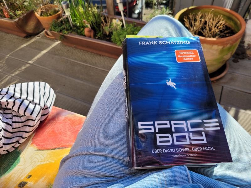 Buch Space Boy von Frank Schätzing auf dem Schoß einer Leserin, die auf einer Bank auf einer Terrasse sitzt. Im Hintergrund Blumenkübel