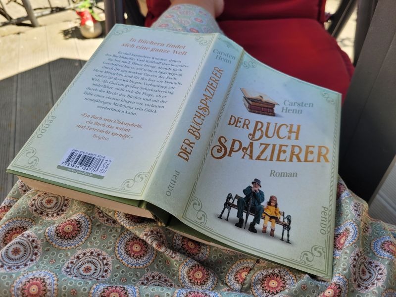 Cover von "Der Buchspazierer" von Carsten Henn aufgeschlagen auf einem Schoß