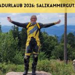 Frau steht in gelbschwarzer Motorradkombi mit ausgebreiteten Armen vor Panorama im Nordschwarzwald. Darüber der Text: Motorradurlaub 2026: Salzkammergut/Kärnten