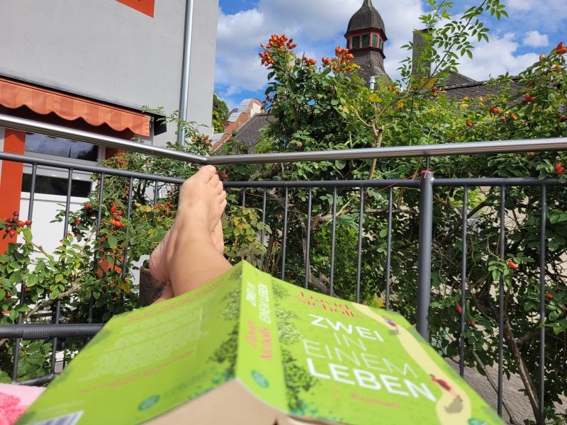 Taschenbuch "Zwei in einem Leben" liegt mit dem Text nach unten auf den Beinen einer auf der Terrasse liegenden Frau. Im Hintergrund blauer Himmel und eine Hagebuttenhecke